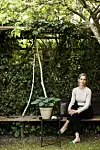 Kvinde sidder på træbænk i grøn have med klatreplanter og lille bord med potteplante.