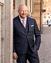 Dennis Lavesson i kritstrecksrandig kostym. Han står lutad mot en husvägg och ler mot kameran.