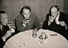 Tre män i kostym sitter vid runt bord och dricker kaffe från fat.