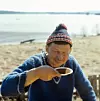 Äldre man i blå tröja dricker kaffe från fat utomhus nära vatten.