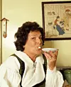 Kvinna i traditionell klädsel dricker kaffe från ett fat och håller en sockerbit i munnen.