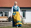 Kvinna i blå och gul svensk folkdräkt och vit hijab står på gräsmatta framför ett vitt hus.