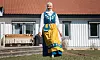 Kvinna i svensk folkdräkt och hijab står barfota på gräsmatta framför ett vitt hus.