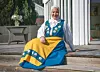 Kvinna i blå och gul Sverigedräkt med hijab sitter på en altan utanför ett hus.
