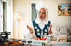 Kvinna i folkdräkt och hijab sitter vid ett dukat bord och gestikulerar i ett ljust vardagsrum.