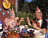 Två personer i pappershattar äter kräftor och skålar vid ett dukat bord utomhus.