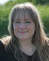 Leende kvinna med långt hår utomhus framför suddig grön natur.