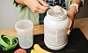 Hand som fyller måttsked med proteinpulver bredvid shaker och bananer på bänk.