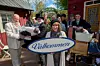 Leende ensemble utomhus håller en stor skylt med texten ”Välkommen” framför röd trähusfasad.