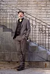 En man i mörk kostym och hatt står framför en stenvägg och en trappa med smidesräcke.