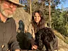 Två personer sitter utomhus på en sten i skogen tillsammans med en svart hund.