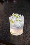 Ljus drink med is, örter och små vita blommor på mörk bordsskiva.