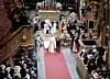 Brudpar och gäster i en kyrka med röd matta, blomsterdekorationer och många åskådare längs gångarna.