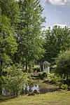 Trädgård med damm, liten vit paviljong och täta gröna träd under en ljus himmel.