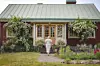 Röd gårdsbyggnad med grön takplåt, blommande urnor vid entrén och en person på gångstigen.