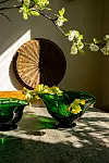 Två gröna glasskålar på ett bord med blommiga kvistar och en rund flätad väggdekoration i bakgrunden.