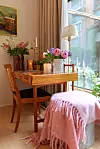 Träbord vid fönster med blomvaser, tända ljus, stol och en rosa pläd över en pall.