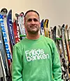 En man i grön Fritidsbanken-tröja står framför en vägg med uppställda skidor och sportutrustning.