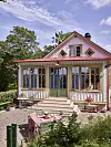 Liten ljus villa med veranda, trädgårdsmöbler och blommande uteplats framför ett trädgårdshus.