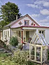 Ljus villa med veranda, små fönster och växthus i förgrunden, omgiven av grönska och blommor.
