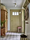 Ljus hall med rutigt golv, beige dörr, träskåp, klädkrokar och en liten pall med rosa kudde.