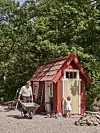 Litet röd-vitt trädgårdshus i en skogsdunge med en skottkärra, en stege och en cykel framför.