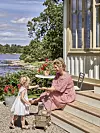 En kvinna i rödvit rutig klänning sitter på en trappa vid ett sommarhus och räcker en sak till ett litet barn.