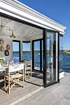 Ljus sjöbod med öppna glasdörrar, dukat bord, stolar och utsikt över en fjord.
