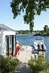 Vit sjöbod vid en träbrygga med en båt, uteplats, växter och fjord i bakgrunden.