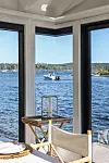 Interiör med stora fönster mot en fjord, en liten båt på vattnet och en ljus stol i förgrunden.