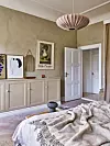 Sovrum med beige kalkmålade väggar, ljusa skåp, tavlor, öppen dörr och en rosa taklampa.