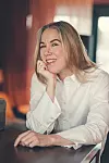 Leende kvinna i vit skjorta sitter vid ett bord med handen mot kinden.
