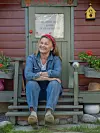 En kvinna sitter på en trappa framför en grön dörr, med blomlådor och en gul fågelholk bredvid.