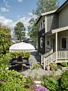 En grusgång leder till en uteplats med vitt parasoll och stolar intill ett grått hus med veranda.