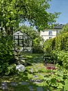 Grönskande trädgård med växthus, stenlagd gång, bänkar och ett hus i bakgrunden.