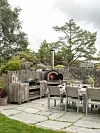 Skifferplattor på grus vid ett bord med stolar, grill och en stenmur i en trädgård.