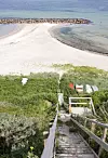 Trätrappa med räcken ner genom tät grönska till en ljus sandstrand och lugnt hav.