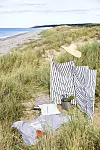 Strandgräs, en randig tygpanel, en stråhatt och flera pysselsaker ligger på sanddyner vid havet.