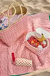 Randig strandväska, kylväska med frukt och dryck samt en rosa filt på sanden.