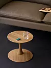 Litet runt träbord framför en grå soffa med ett glas och ett grönt föremål på skivan.