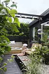 Uteplats med trädeck, soffa, svart träpanel, hängande lampor och tät plantering i förgrunden.