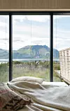 Sovrum med säng framför stora fönster som vetter mot fjäll, vatten och grönskande kustlandskap.