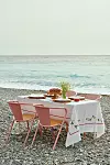 Ett bord dukat på en stenstrand med rosa stolar, vit duk, fat och blommor framför havet.