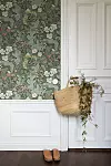 Grön tapet med blommönster, vit panelvägg, korgväska med blommor och ett par skor på golvet.