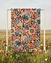En färgstark blom- och fågelmönstrad textil hänger i en ram på en äng med vildblommor.