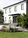 Vit tvåvåningsvilla med trappa, uteplats och grönskande trädgård framför fasaden.