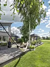 Modernt hus med uterum, grusgång, stora krukor och en hängmatta under ett björkträd vid en gräsmatta.