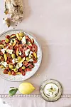 Tallrik med tomatsallad, oliver och mozzarella bredvid citron, bröd och en liten skål med dressing.
