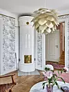 Ljus hall med vit kakelugn, blommig tapet, trägolv och stor pendellampa.