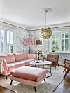 Vardagsrum med rosa soffa, rund soffbord, fotpall, blommig tapet och stora fönster.